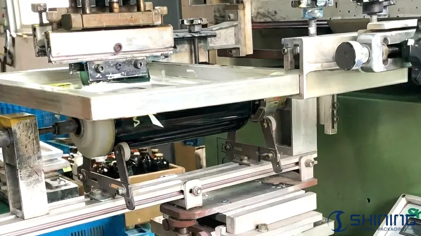 Silk screen printing machine applying thick ink onto aluminum bottles through mesh stencil for bold designs